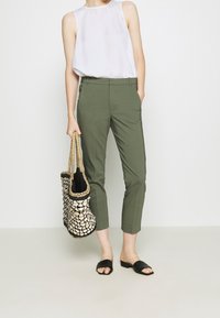 Mujer vestida con una blusa sin mangas blanca y pantalones recortados verdes oliva, sosteniendo un bolso de mimbre y usando sandalias negras tipo slide.