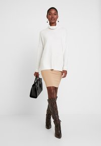 Hvit genser med cowl-nekk, beige bodycon skjørt og brune støvler med snakeskin til kneet. Svart håndveske i hånden, enkle øredobber som aksenter.