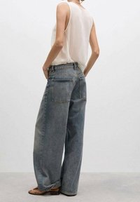 Osoba ubrana w białą bluzkę bez rękawów i luźne, wyblakłe niebieskie dżinsy, z rękami w kieszeniach z tyłu, stojąca na szarym podłodze.