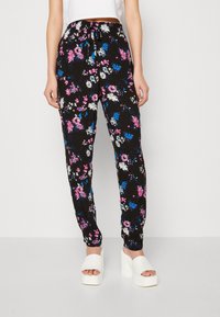 Pantalones florales negros con flores rosas, azules y blancas; cinturilla elástica con un cordón; tobillos acampanados; combinados con zapatos de plataforma blancos.