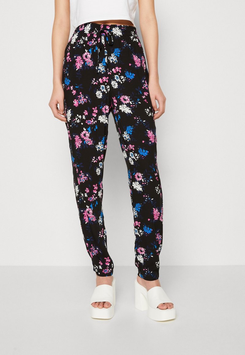 Pantalones florales negros con flores rosas, azules y blancas; cinturilla elástica con un cordón; tobillos acampanados; combinados con zapatos de plataforma blancos.