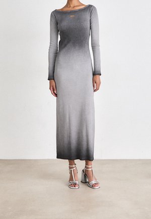Femme portant une robe longue à manches longues, dégradé gris, avec une petite étiquette sur la poitrine et des sandales argentées à talons hauts.