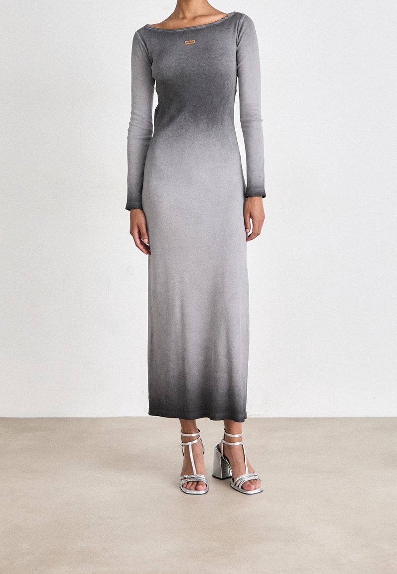 Femme portant une robe longue à manches longues, dégradé gris, avec une petite étiquette sur la poitrine et des sandales argentées à talons hauts.