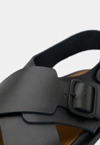 Μαύρο δερμάτινο σανδάλι με φαρδιές τιράντες, ρυθμιζόμενο μεταλλικό κούμπωμα και αντίθετο ταμπά πάτο. Ομαλή υφή και ανοιχτός σχεδιασμός.