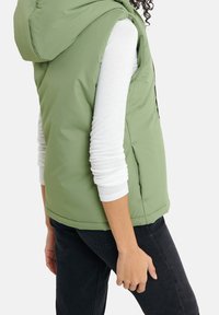 Grön pufferväst med huva, med en slät textur, sidofickor och en figursydd design. Bärs över en vit långärmad skjorta och svarta byxor.
