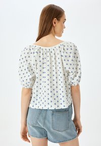 Witte blouse met een blauw driehoekpatroon, opgeblazen mouwen en een brede halslijn. Gecombineerd met hoge denim shorts.