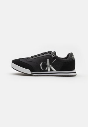 Sneaker noire avec une tige en tissu, des accents en daim, une semelle blanche et un grand logo "CK" argenté sur le côté. Comprend des lacets noirs et des coutures.