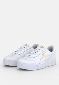 Baskets athlétiques blanches avec extérieur en cuir texturé, bout rond et semelle plate. Présentent des accents de logo dorés et des panneaux latéraux contrastés.