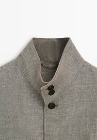 Camicia con colletto alto in tessuto grigio strutturato con tre bottoni neri visibili sulla patta frontale.