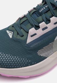 Zapatilla de running con parte superior de malla texturizada en azul-verde, suela rosa claro y acentos grises. Cuenta con una lengüeta acolchada y puntera reforzada.