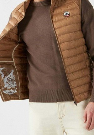 Homme portant un pull marron et une veste matelassée beige avec logo, tenant la veste ouverte pour montrer le motif imprimé de la doublure intérieure.