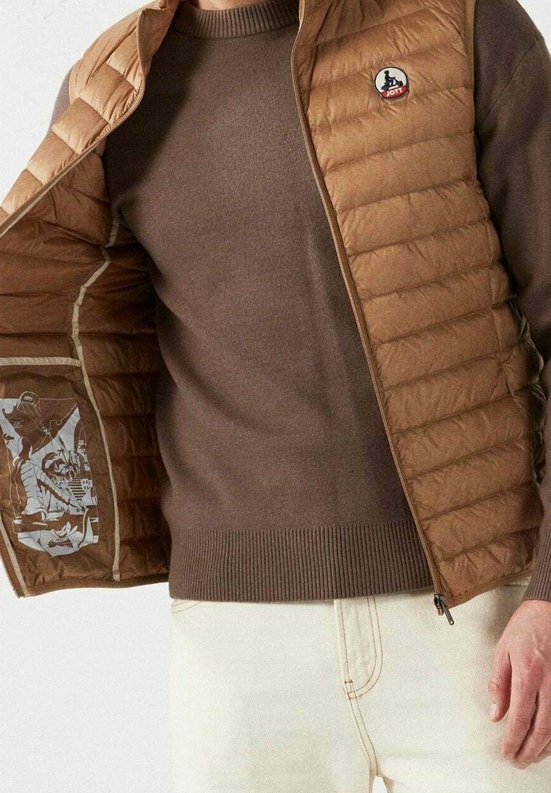 Homme portant un pull marron et une veste matelassée beige avec logo, tenant la veste ouverte pour montrer le motif imprimé de la doublure intérieure.