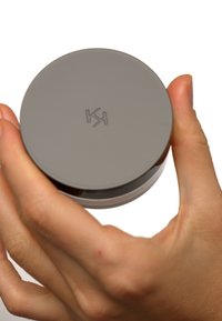 Grå rund beholder holdt i en hånd, glatt overflate med preget logo på toppen, plastmateriale, litt gjennomsiktig bunn.