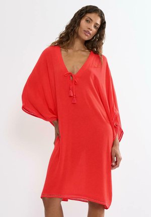 Frau mit langen lockigen Haaren, die ein lockeres rotes Kleid mit V-Ausschnitt und Quastenbändern trägt, posiert mit einer Hand in der Hüfte vor einem einfarbigen Hintergrund.