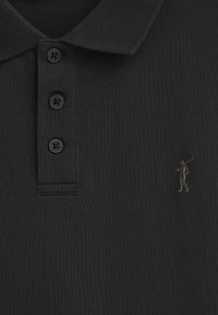 Svart pikétröja med en strukturerad tyg, fyra svarta knappar och en subtil, broderad figur i en ljusare nyans på vänster bröst.