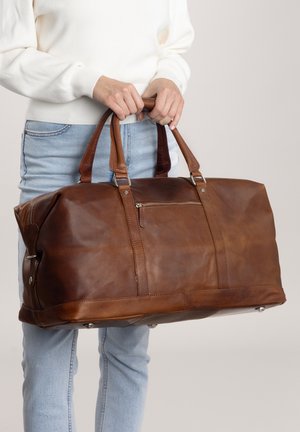 Sac de voyage en cuir marron avec deux poignées, une poche zippée à l'avant, quincaillerie en métal et pieds de protection au fond. Fini lisse et texturé.