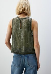 Olijfgroene mouwloze top van textuurstof met pailletten en een sleutelgatdetail op de rug, gecombineerd met lichtblauwe spijkerbroek.