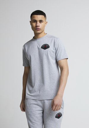 Ung mann iført en matchende lys grå kortermet skjorte og joggebukser med svarte og røde "Zavetti"-logoer på brystet og låret.