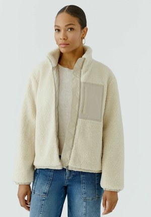 Femme portant une veste zippée texturée couleur crème sur un pull et un jean bleu, debout devant un fond clair uni.