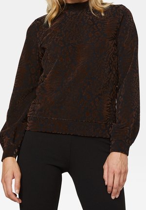 Kvinde iført en mørkebrun og sort langærmet bluse med subtilt dyreprint og sorte bukser, hænder afslappet langs siderne.