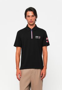Sort polo med korte ærmer, med en frontlynl�ås og USA Sail GP Team-logo på brystet og ærmet. Kontrasterende kravekanter.