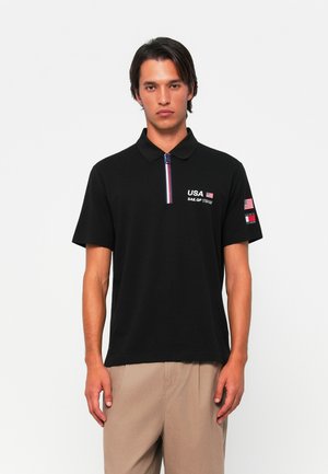 Juodas polo marškinėliai su trumpomis rankovėmis, kuriuose yra priekinis užtrauktukas ir JAV Sail GP komandos logotipas ant krūtinės ir rankovės. Priekinis apykaklės akcentas skiriasi.