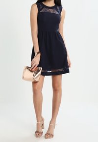 Mörkblå ärmlös klänning med spetsinlägg vid halsringningen och fållen, i kombination med en metallisk roséguldfärgad clutch och sandaler med remmar.