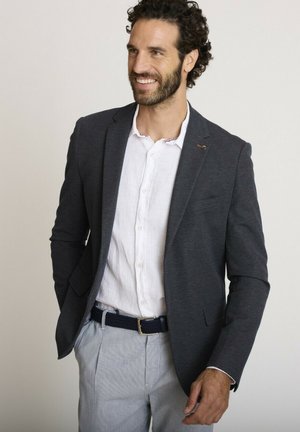 Homme aux cheveux bouclés et foncés, barbu, portant un blazer sombre, une chemise blanche, un pantalon rayé, souriant, debout devant un fond uni.