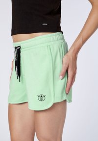 Detailní záběr na osobu v světle zelených šortkách na šňůrku s černým logem, které jsou v kombinaci s černým zkráceným topem, s rukou položenou na boku.