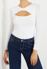 Woman wearing white long-sleeve top with twist front cutout paired with dark blue high-waisted jeans, showing torso and hands.