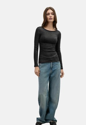 Donna in piedi che indossa un top nero aderente a maniche lunghe e jeans larghi azzurro chiaro su uno sfondo chiaro e uniforme.