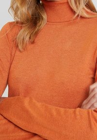 Pomarańczowy sweter z golfem, z prążkowanym kołnierzem i długimi rękawami; wykonany z miękkiej, fakturowanej dzianiny. Zbliżenie na górną część ciała.