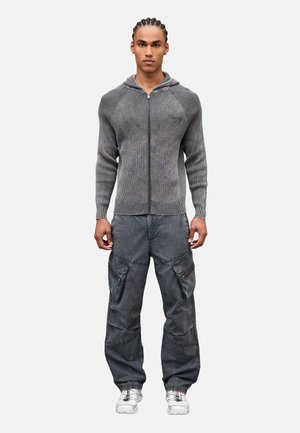 Mann mit geflochtenen Haaren, der einen grauen Zip-Up-Kapuzenpullover, dunkle Cargo-Hosen und weiße Turnschuhe trägt und mit dem Gesicht nach vorne vor einem einfarbigen Hintergrund steht.