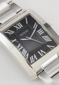 Montre en acier inoxydable argenté avec un cadran rectangulaire noir, des chiffres romains argentés et deux aiguilles argentées sur un bracelet en métal maillé.