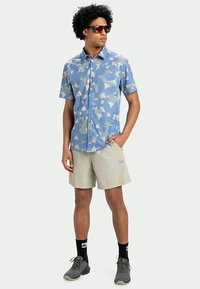 Kortärmad, ljusblå skjorta med knappar som har ett mönster av abstrakta former och blommiga detaljer, ihop med beige shorts och grå sneakers.