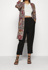 Kobieta w czarnych spódniczkach, białej bezrękawnikowej bluzce, długim kardiganie w kwiatowy wzór i czarnych sandałach na obcasach, stoi z ręką w kieszeni.