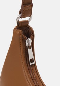 Gros plan de la sangle en cuir marron d'un sac à main attachée avec un anneau argenté et un curseur de fermeture éclair argenté visible sur une doublure en tissu marron.
