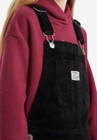 Černé kordové kšandy se stříbrným kováním a nastavovacími popruhy, nošené přes vínový mikina. Obsahuje přední kapsu s etiketou.