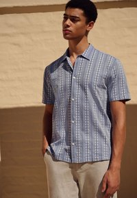 Chemise à manches courtes, boutonnée, de couleur bleu clair avec des rayures blanches verticales et des motifs floraux, fabriquée dans un tissu léger.