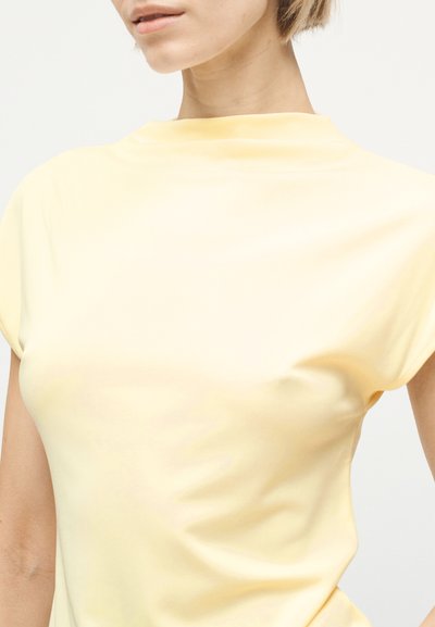 Γυναίκα που φοράει αμάνικη γκρι κίτρινη μπλούζα με υψηλό λαιμόκοψη, εμφανίζεται από τους ώμους έως το μέσο του κορμού σε απλό φόντο.