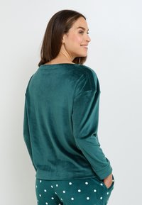 Top turchese a maniche lunghe realizzato in morbido tessuto vellutato. Presenta un ampio scollo e una vestibilità rilassata, abbinato a un pantalone da casa verde a pois.