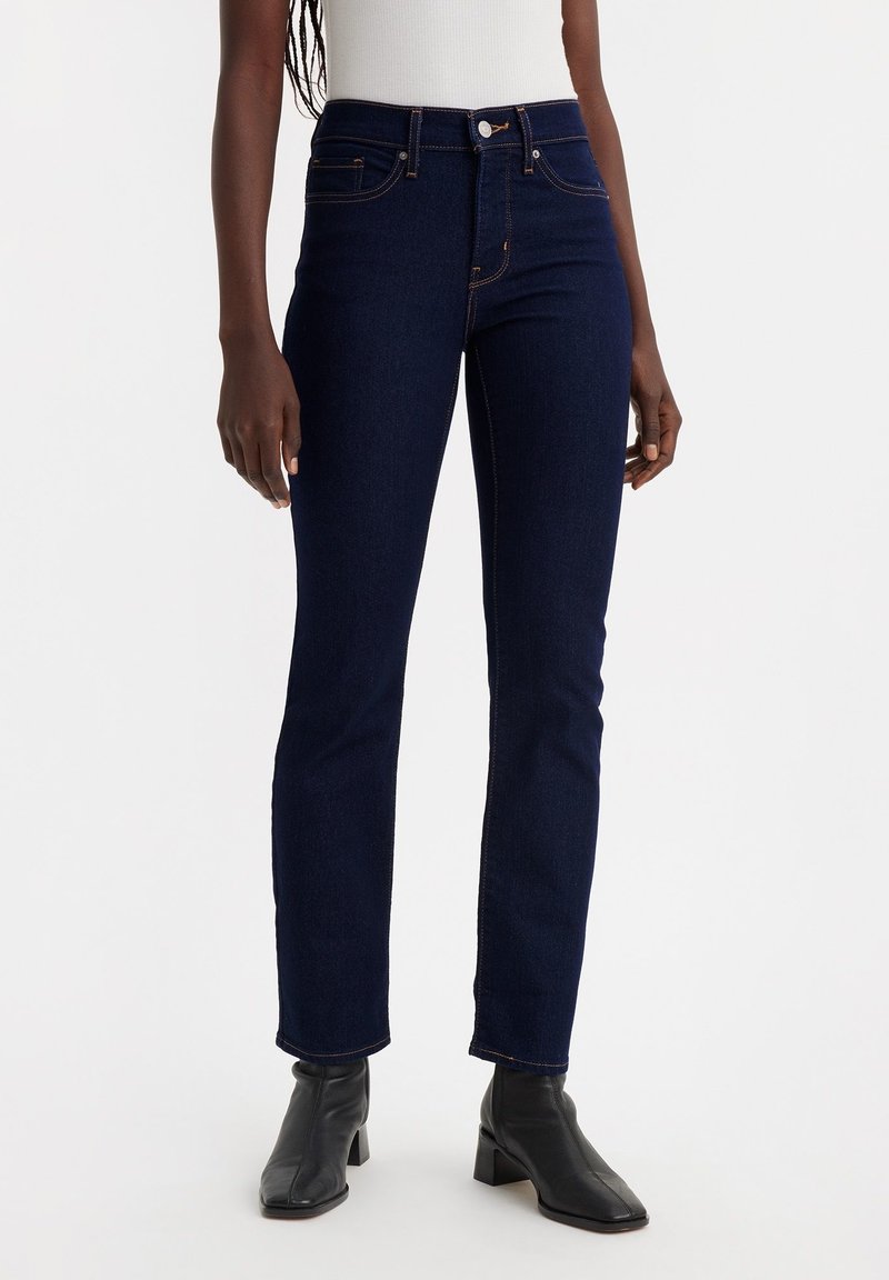 Femme portant un jean droit bleu foncé, un haut blanc sans manches et des bottines noires à bout carré sur un fond clair uni.