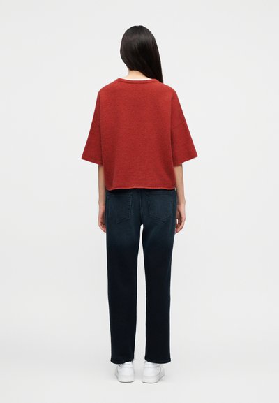 Γυναίκα με μακριά σκουρόχρωμα μαλλιά στέκεται γυρισμένη με την πλάτη, φοράει ένα φαρδύ κόκκινο τοπ, σκούρα τζιν με ίσιο κόψιμο και άσπρα αθλητικά παπούτσια πάνω σε ουδέτερο φόντο.