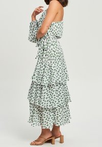 Vestido blanco con estampado floral verde, diseño en capas, cinturilla ajustable con lazo y mangas largas. Combinado con sandalias de tacón cuadrado de color marrón claro.