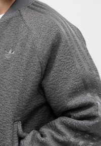 Mangă de jachetă din fleece texturat gri, cu guler ribbed, logo Adidas pe piept și trei dungi tonale pe umăr.