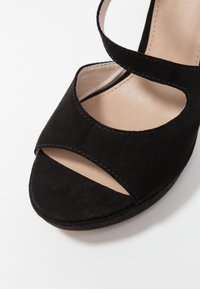 Svart mocka högklackad sandal med öppen tå, försedd med korslagda remmar och en slät beige innersula. Designen har subtila sömdetaljer.