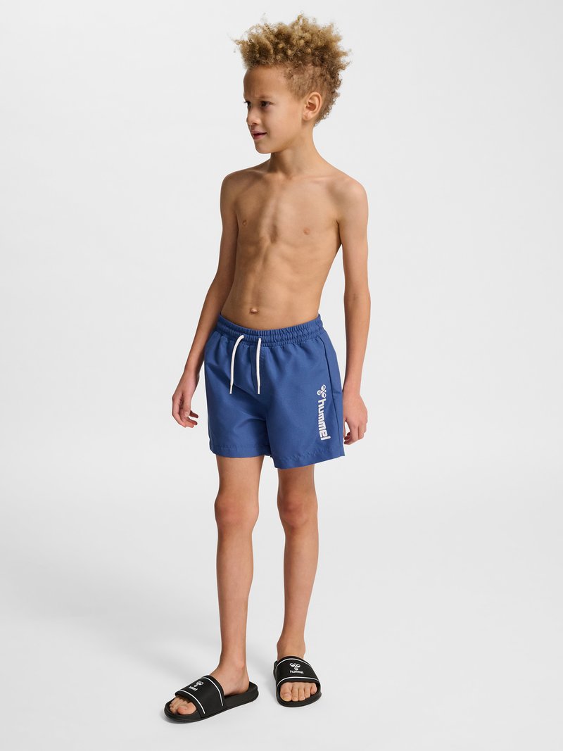 Junge mit lockigem Haar, der blaue Badeshorts und schwarze Slides trägt, steht und schaut zur Seite auf weißem Hintergrund.