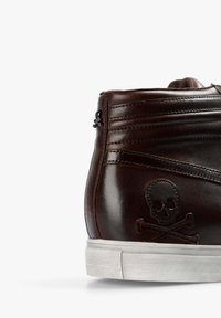 Zapatilla alta marrón hecha de cuero brillante, con un diseño de calavera y huesos cruzados, paneles texturizados y una suela de goma blanca.