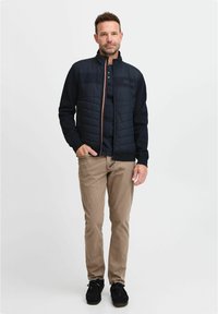 Marineblaues, gesteppte Jacke mit hohem Kragen, Frontreißverschluss und kontrastierenden orangefarbenen Akzenten, kombiniert mit beigen Hosen und schwarzem Schuhwerk.