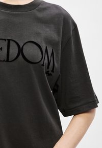 Σκούρο γκρι βαμβακερό t-shirt με κοντά μανίκια και στρογγυλή λαιμόκοψη. Το μπροστινό μέρος έχει έντονα μαύρα γράμματα που λένε "ΕΛΕΥΘΕΡΙΑ".